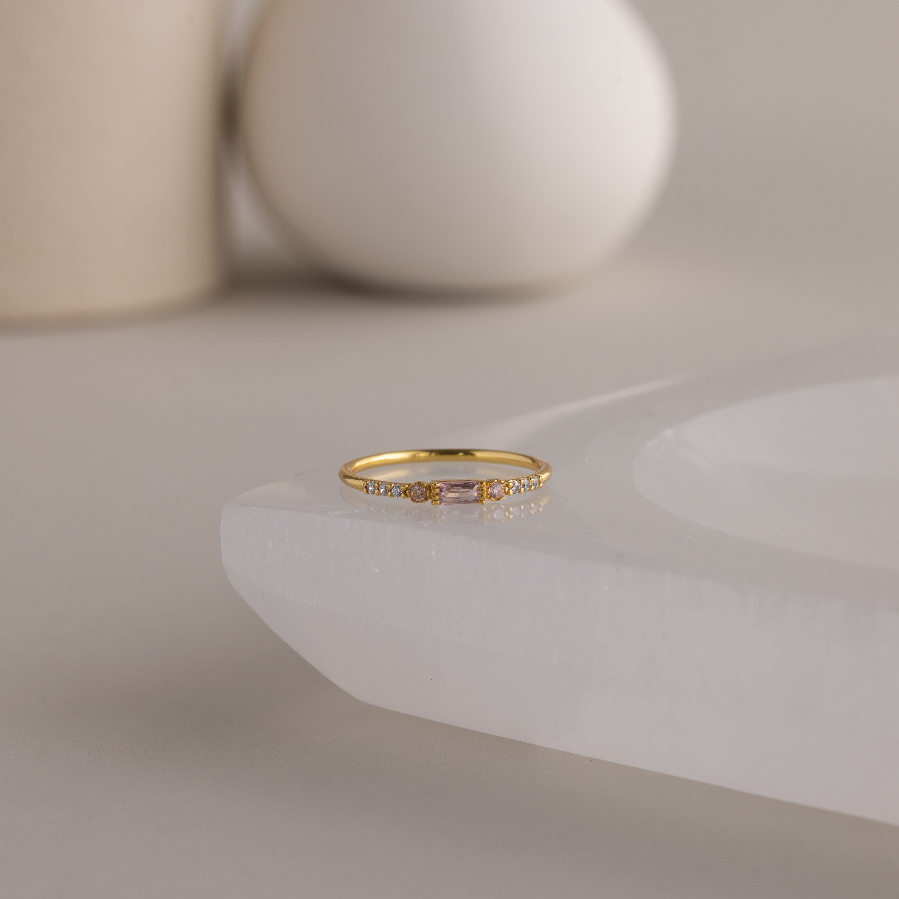 A single dainty gold birthstone ring with a rectangular pink tourmaline gemstone and small white accent stones rests on a curved white stone surface with minimalist background decor.