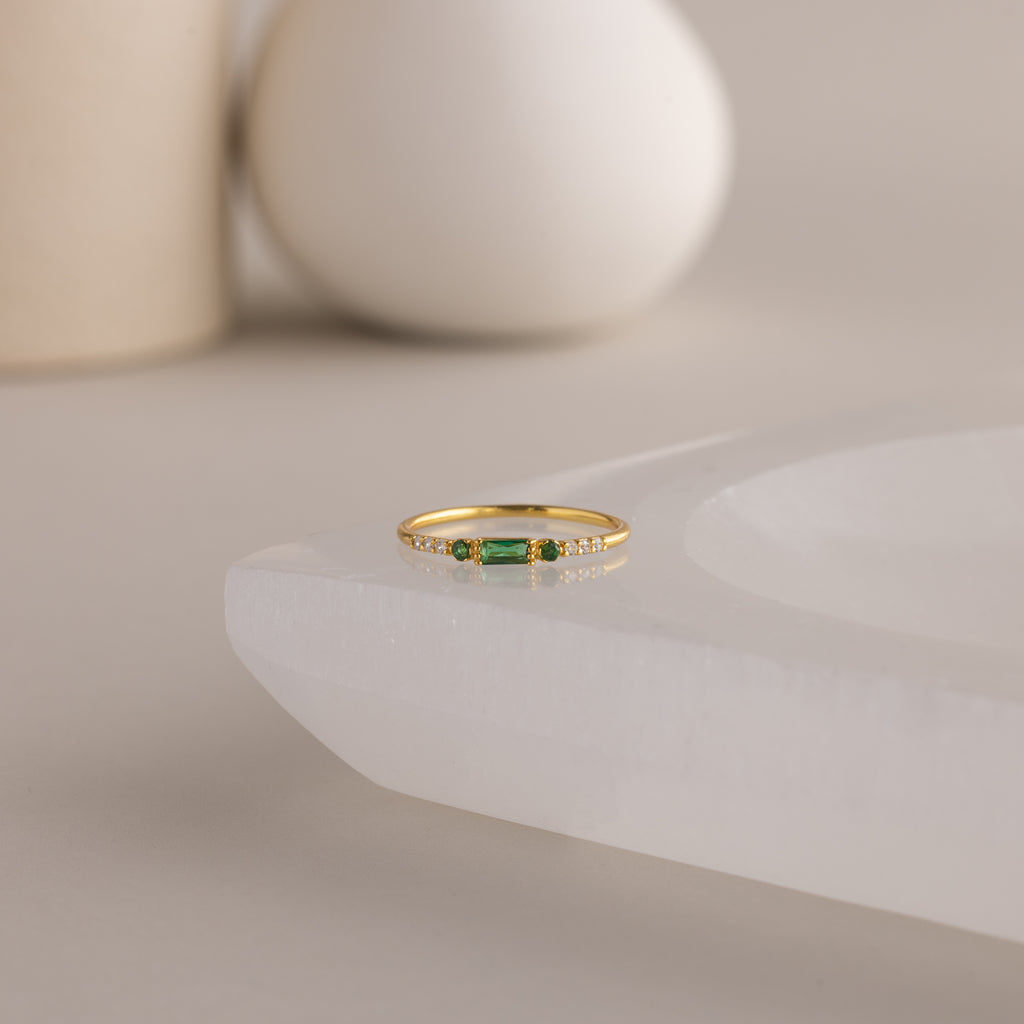 A single dainty gold birthstone ring with a rectangular emerald green gemstone and small white accent stones rests on a curved white stone surface with minimalist background decor.