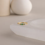 A single dainty gold birthstone ring with a rectangular emerald green gemstone and small white accent stones rests on a curved white stone surface with minimalist background decor.