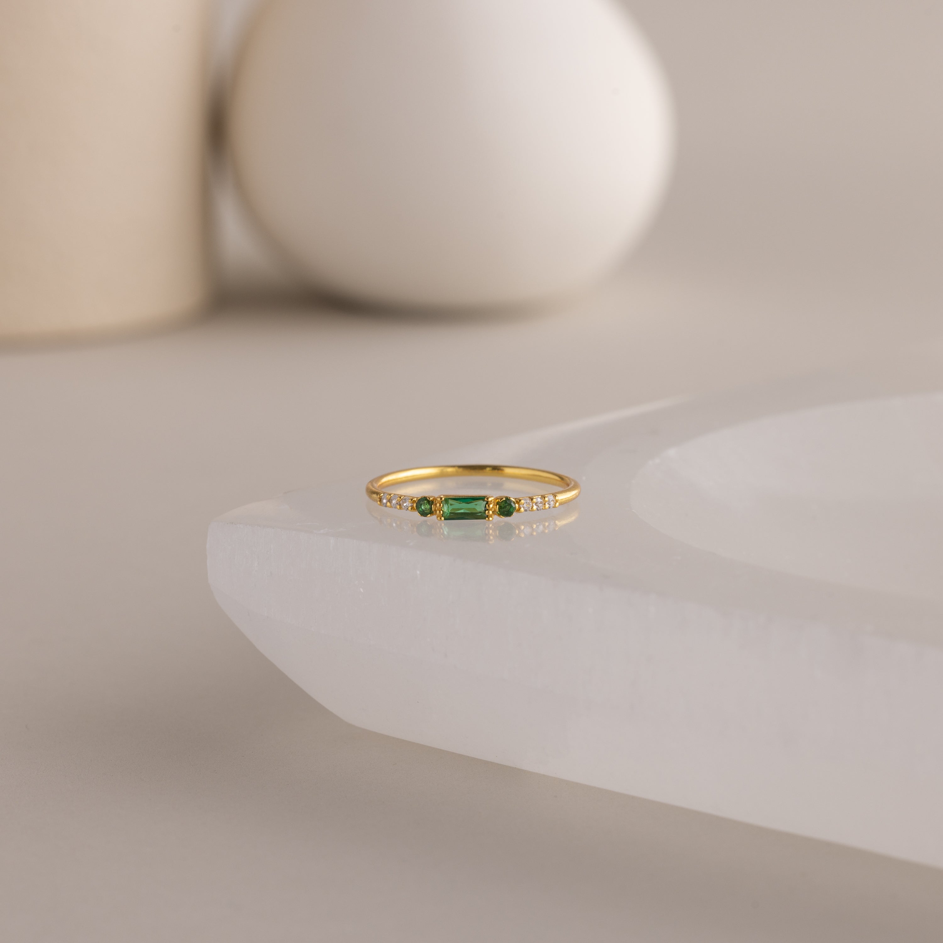 A single dainty gold birthstone ring with a rectangular emerald green gemstone and small white accent stones rests on a curved white stone surface with minimalist background decor.