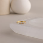 A single dainty gold birthstone ring with a rectangular light alexandrite gemstone and small white accent stones rests on a curved white stone surface with minimalist background decor.