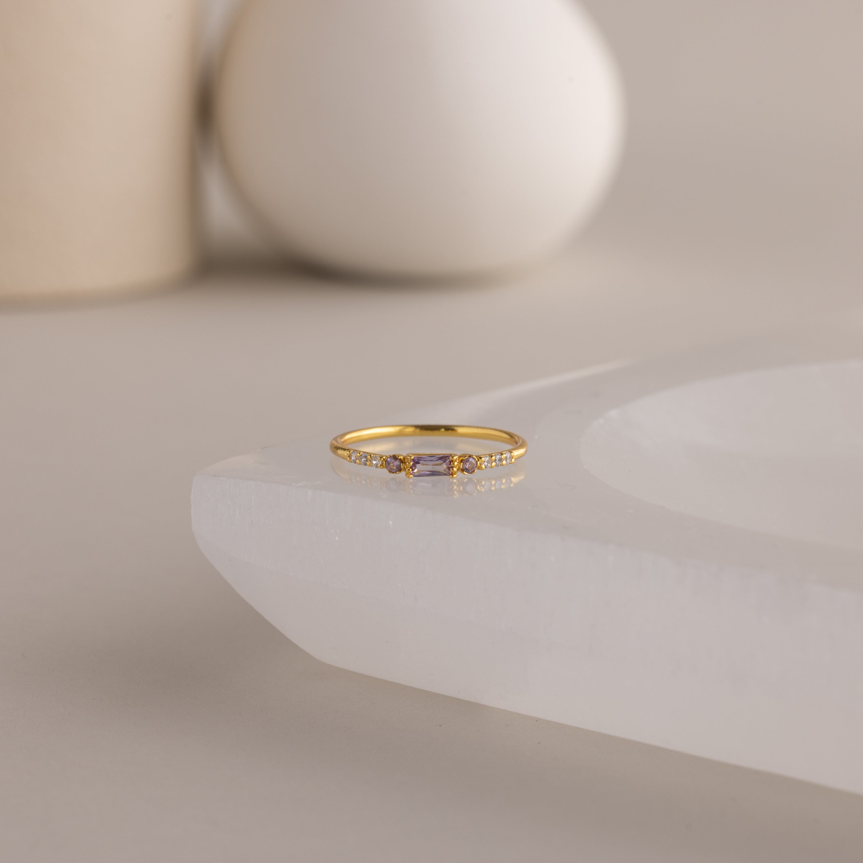 A single dainty gold birthstone ring with a rectangular light alexandrite gemstone and small white accent stones rests on a curved white stone surface with minimalist background decor.