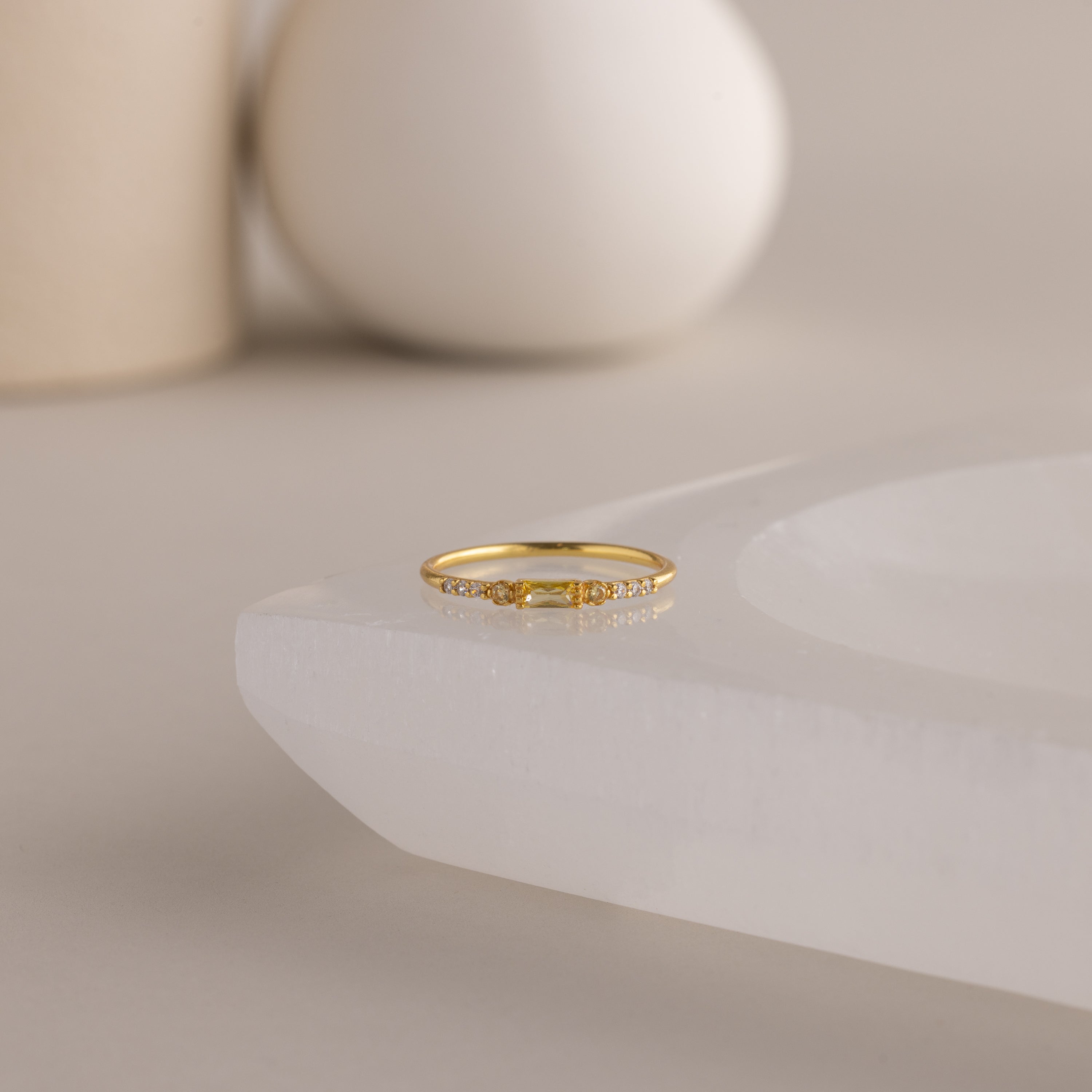 A single dainty gold birthstone ring with a rectangular yellow topaz gemstone and small white accent stones rests on a curved white stone surface with minimalist background decor.