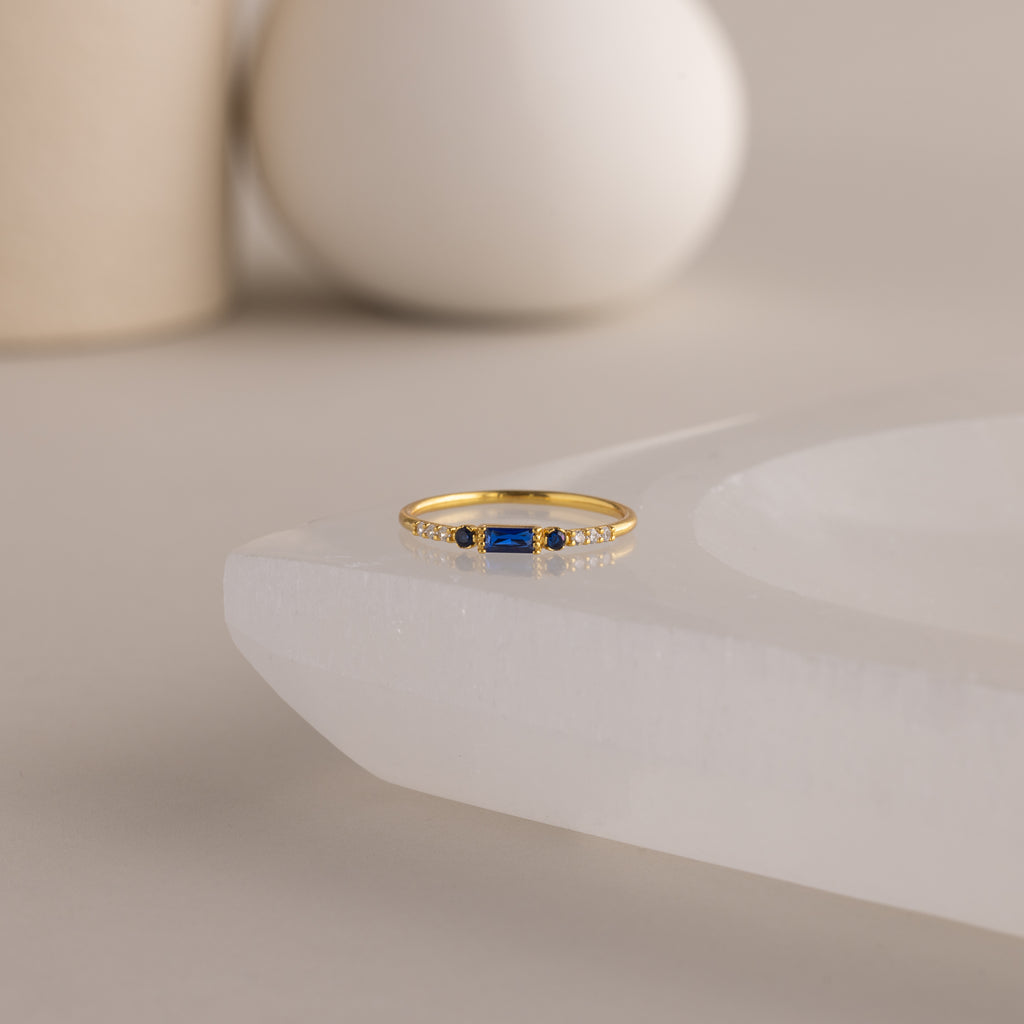 A single dainty gold birthstone ring with a rectangular sapphire gemstone and small white accent stones rests on a curved white stone surface with minimalist background decor.