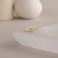 A single dainty gold birthstone ring with a rectangular diamond gemstone and small white accent stones rests on a curved white stone surface with minimalist background decor.