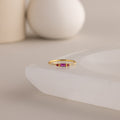 A single dainty gold birthstone ring with a rectangular ruby gemstone and small white accent stones rests on a curved white stone surface with minimalist background decor.