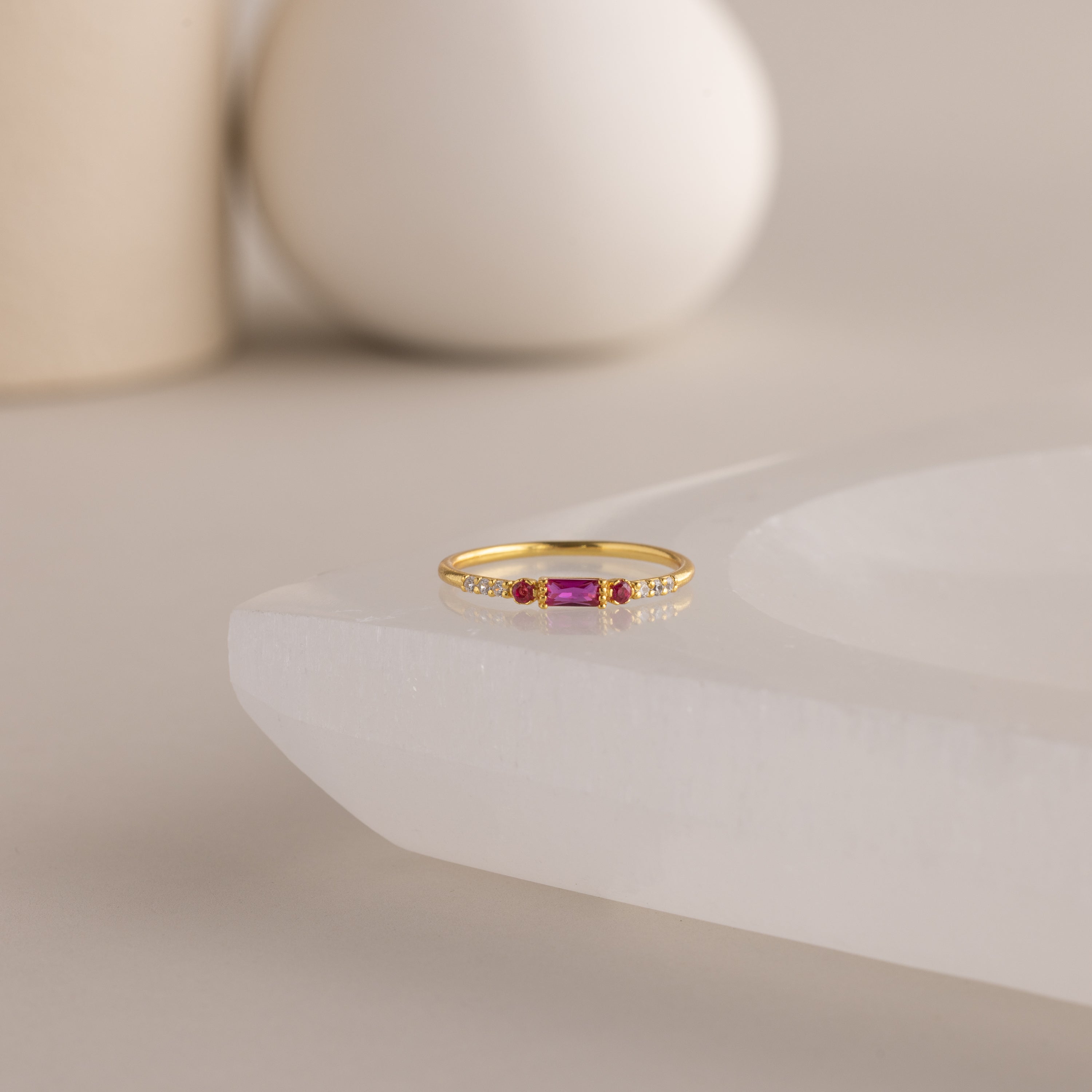 A single dainty gold birthstone ring with a rectangular ruby gemstone and small white accent stones rests on a curved white stone surface with minimalist background decor.