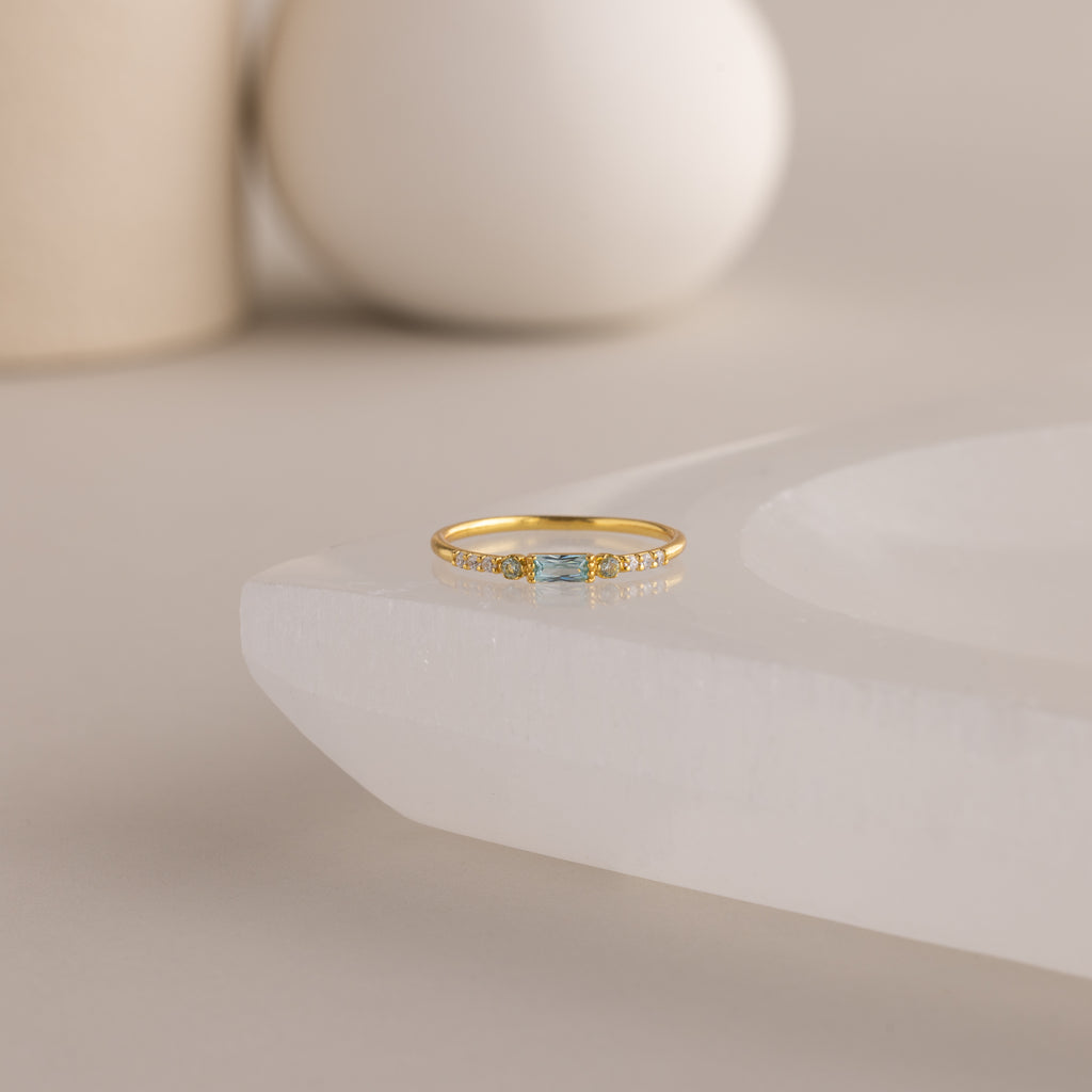 A single dainty gold birthstone ring with a rectangular aquamarine gemstone and small white accent stones rests on a curved white stone surface with minimalist background decor.
