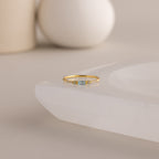 A single dainty gold birthstone ring with a rectangular aquamarine gemstone and small white accent stones rests on a curved white stone surface with minimalist background decor.