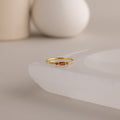 A single dainty gold birthstone ring with a rectangular red gemstone and small white accent stones rests on a curved white stone surface with minimalist background decor.