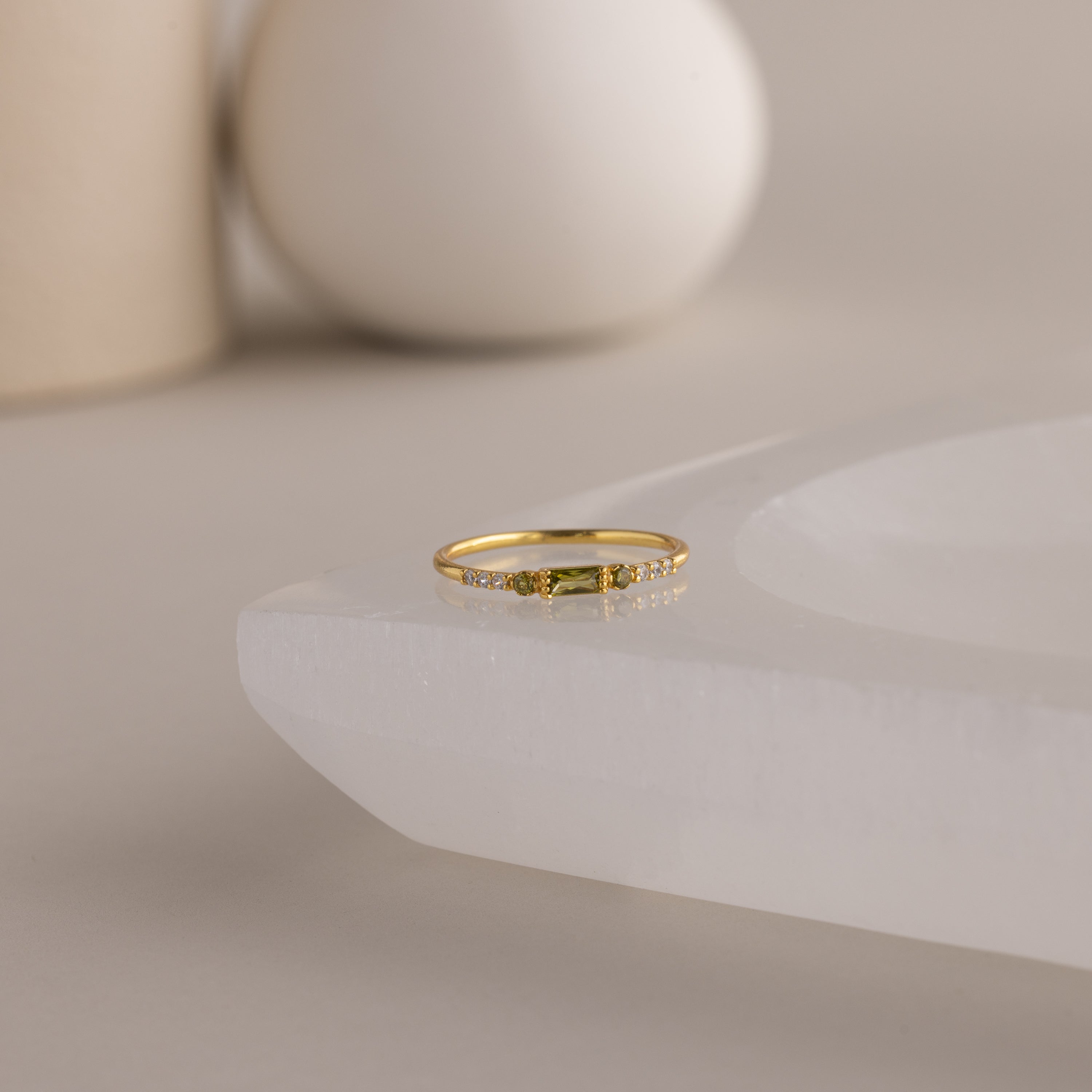 A single dainty gold birthstone ring with a rectangular peridot gemstone and small white accent stones rests on a curved white stone surface with minimalist background decor.