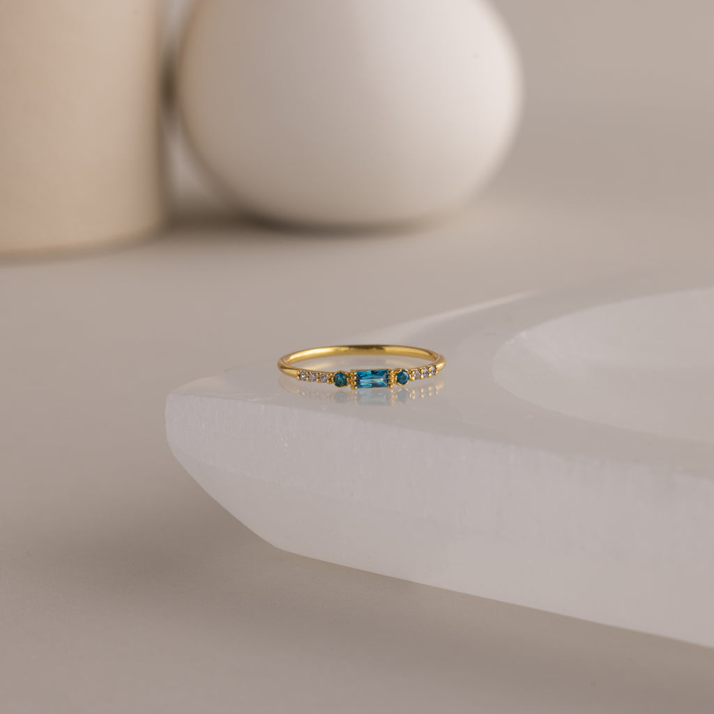 A single dainty gold birthstone ring with a rectangular blue zircon gemstone and small white accent stones rests on a curved white stone surface with minimalist background decor.