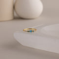 A single dainty gold birthstone ring with a rectangular blue zircon gemstone and small white accent stones rests on a curved white stone surface with minimalist background decor.