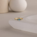 A single dainty gold birthstone ring with a rectangular blue zircon gemstone and small white accent stones rests on a curved white stone surface with minimalist background decor.