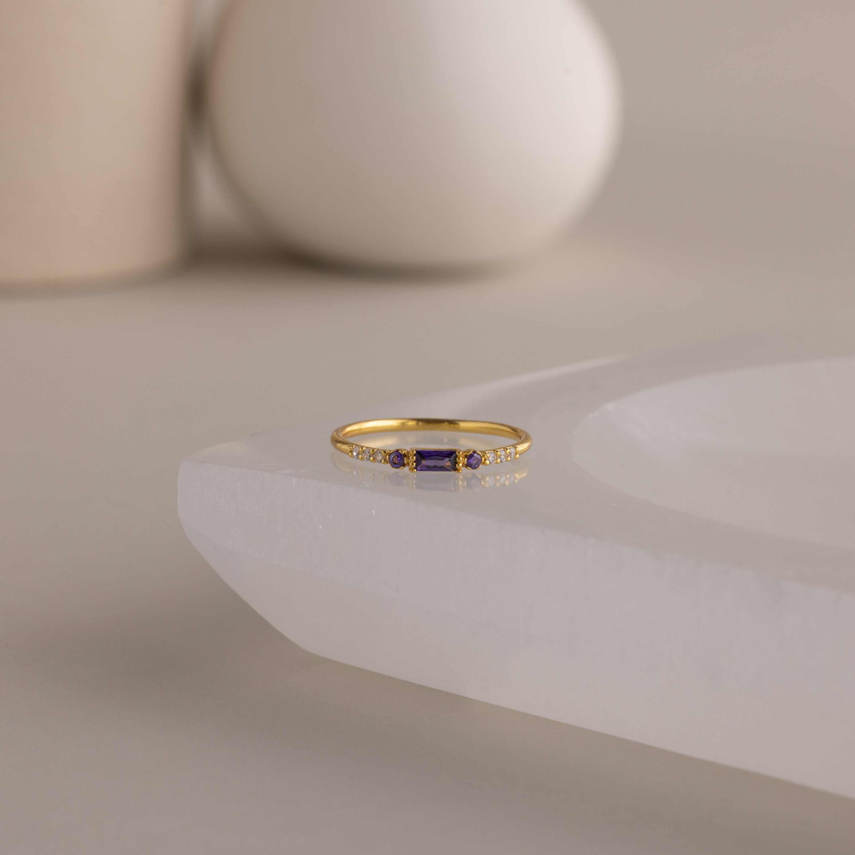 A single dainty gold birthstone ring with a rectangular amethyst gemstone and small white accent stones rests on a curved white stone surface with minimalist background decor.