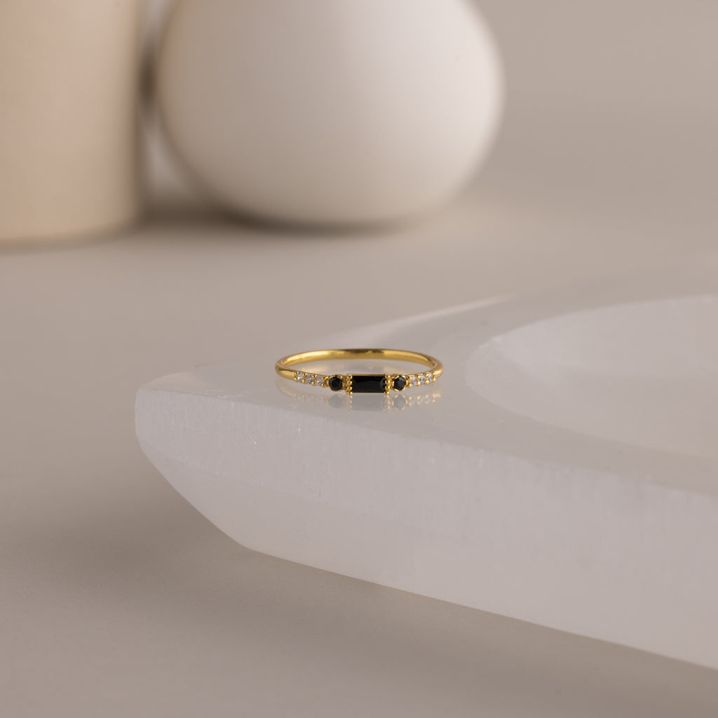 A single dainty gold birthstone ring with a rectangular onyx gemstone and small white accent stones rests on a curved white stone surface with minimalist background decor.