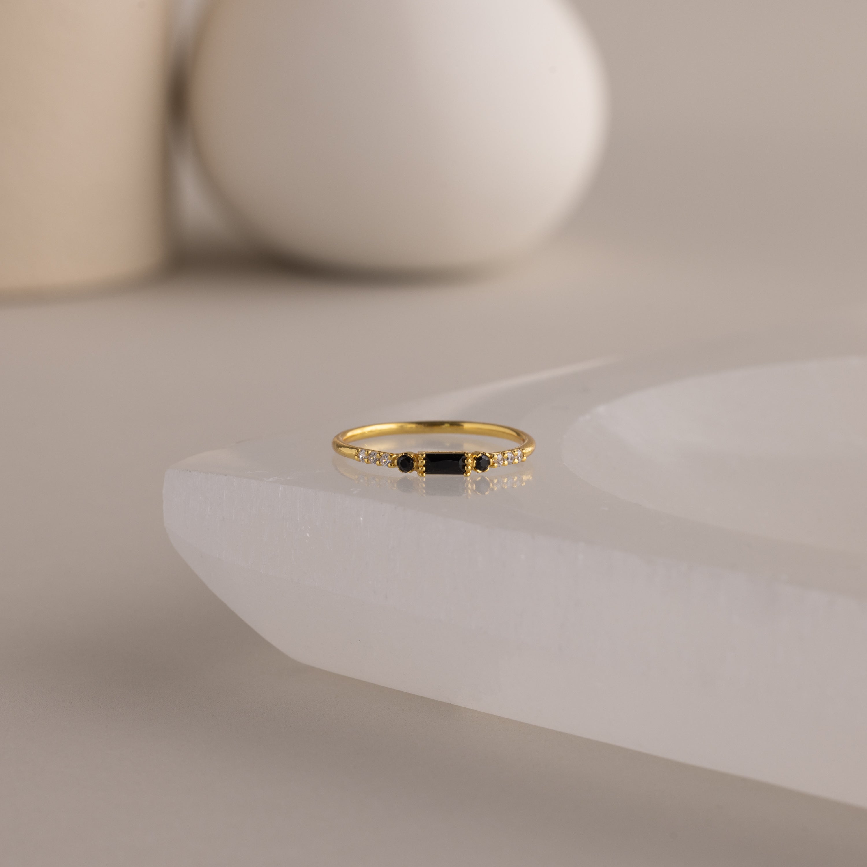 A single dainty gold birthstone ring with a rectangular onyx gemstone and small white accent stones rests on a curved white stone surface with minimalist background decor.