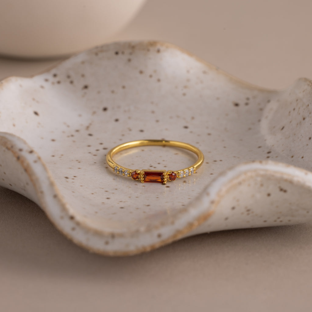A dainty gold ring with a rectangular garnet baguette-cut gemstone flanked by small clear stones, displayed elegantly in a speckled ceramic dish with soft lighting.