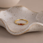 A dainty gold ring with a rectangular garnet baguette-cut gemstone flanked by small clear stones, displayed elegantly in a speckled ceramic dish with soft lighting.