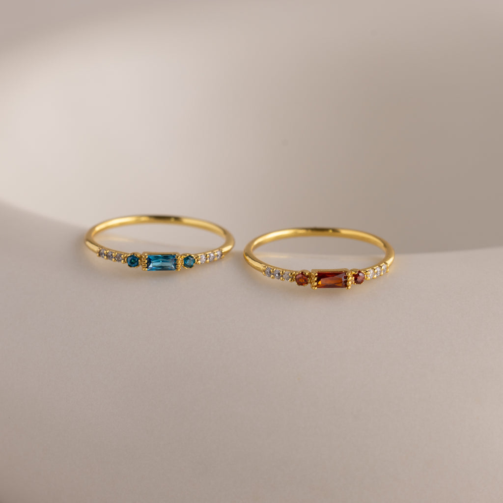 Two delicate gold birthstone rings side by side on a white stone slab—one with a garnet baguette gemstone, the other with a vibrant blue zircon baguette stone—each accented with petite clear stones along the band.