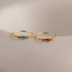 Two delicate gold birthstone rings side by side on a white stone slab—one with a garnet baguette gemstone, the other with a vibrant blue zircon baguette stone—each accented with petite clear stones along the band.