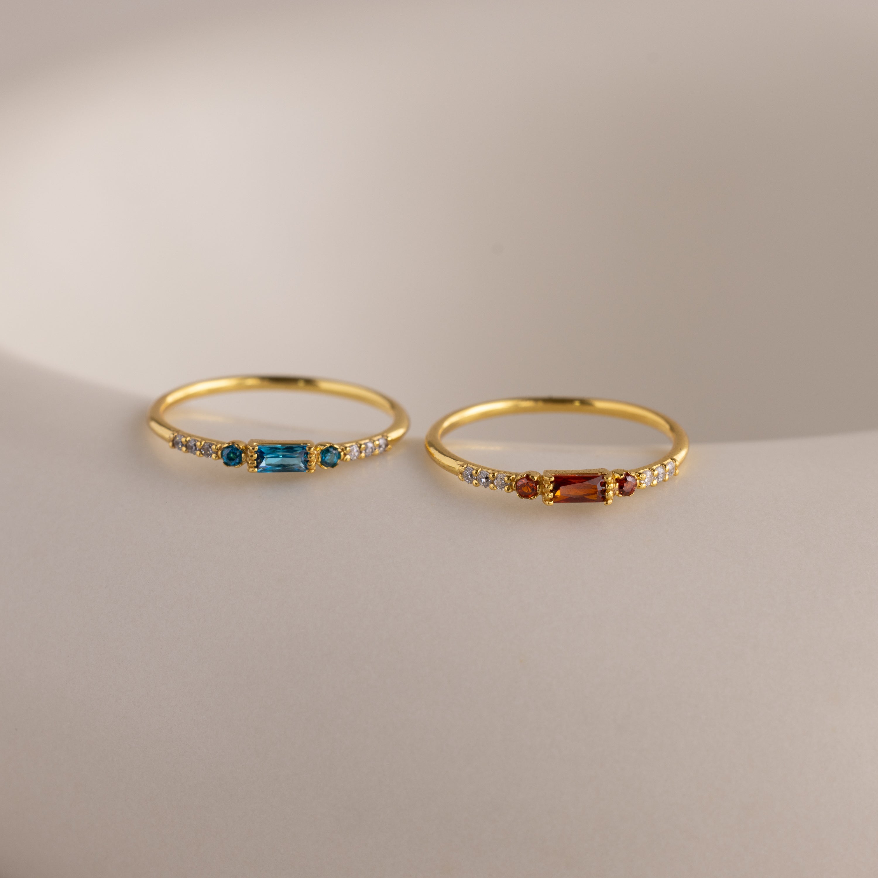 Two delicate gold birthstone rings side by side on a white stone slab—one with a garnet baguette gemstone, the other with a vibrant blue zircon baguette stone—each accented with petite clear stones along the band.