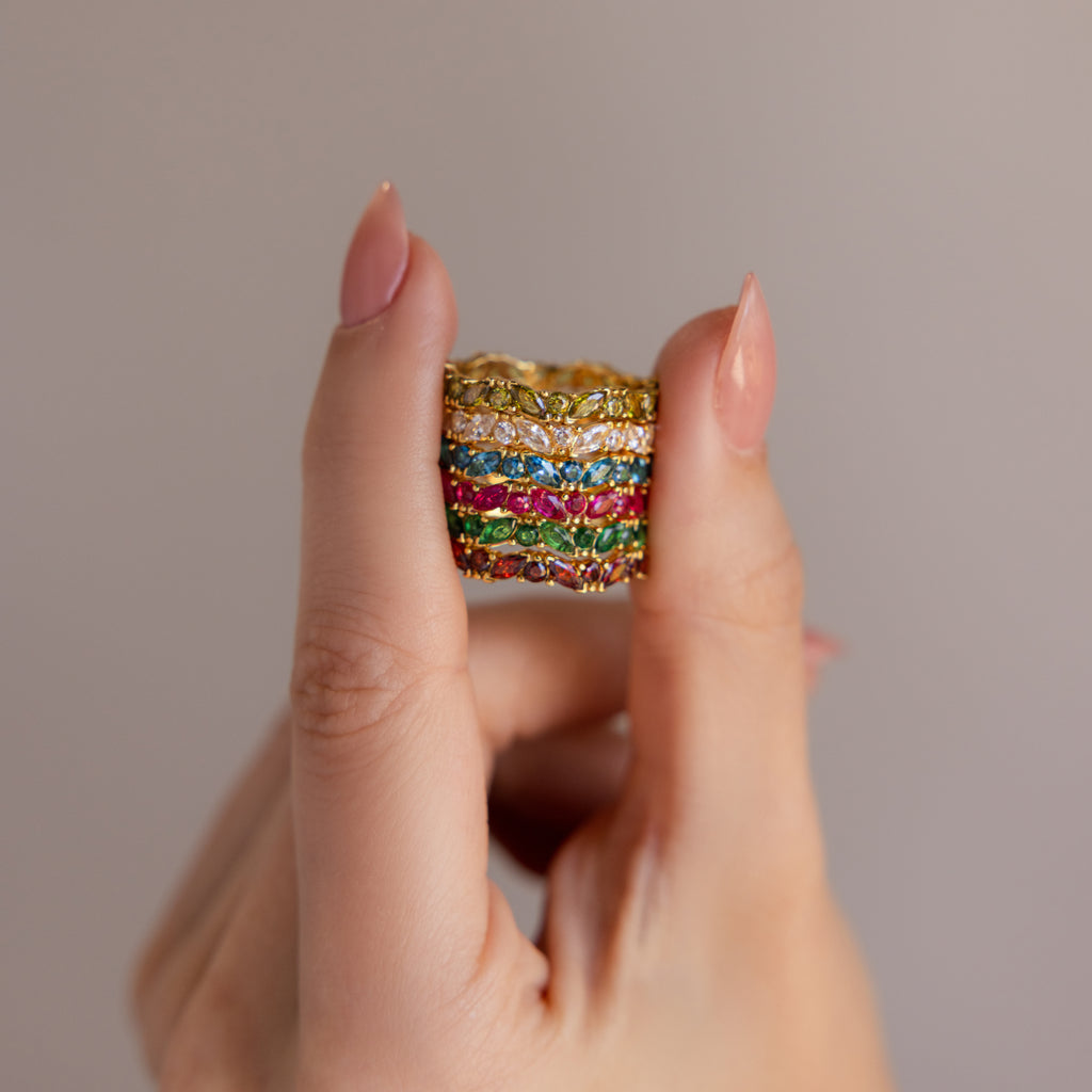 A hand displays stacked gold rings, featuring the Staggered Marquise Diamond Ring alongside a colorful gemstone eternity band, set against a plain background.