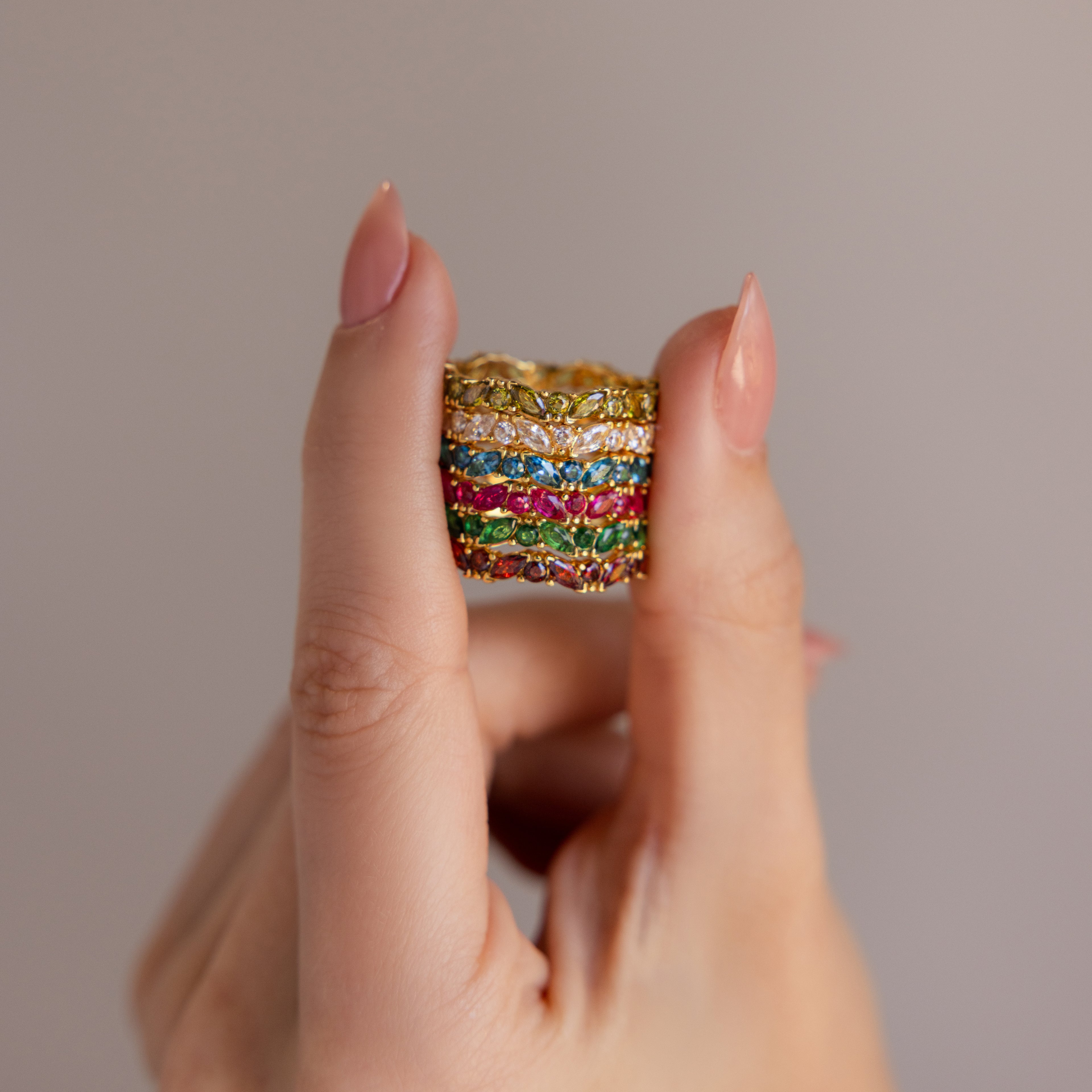 A hand displays stacked gold rings, featuring the Staggered Marquise Diamond Ring alongside a colorful gemstone eternity band, set against a plain background.