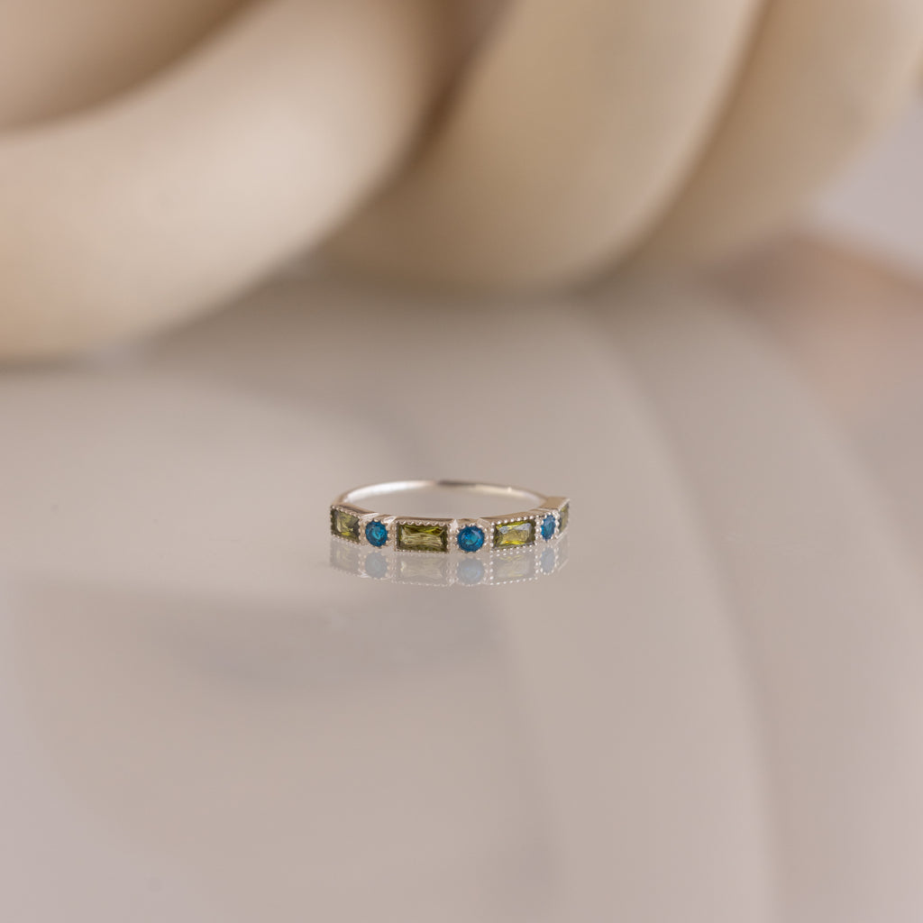 The Mixed Art Deco Birthstone Ring in Sterling Silver features alternating blue and green stones, displayed on a reflective surface against a soft beige background.