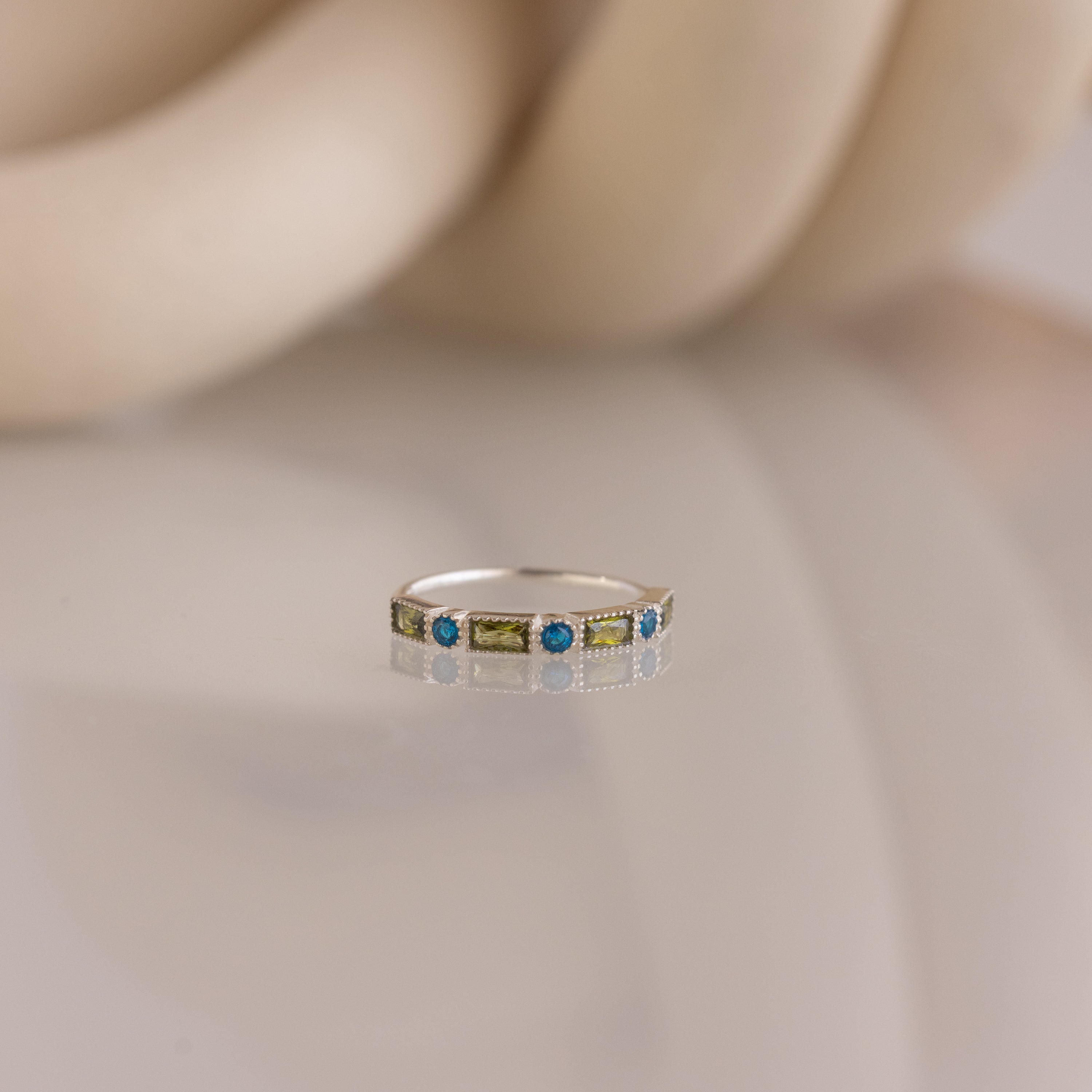 The Mixed Art Deco Birthstone Ring in Sterling Silver features alternating blue and green stones, displayed on a reflective surface against a soft beige background.