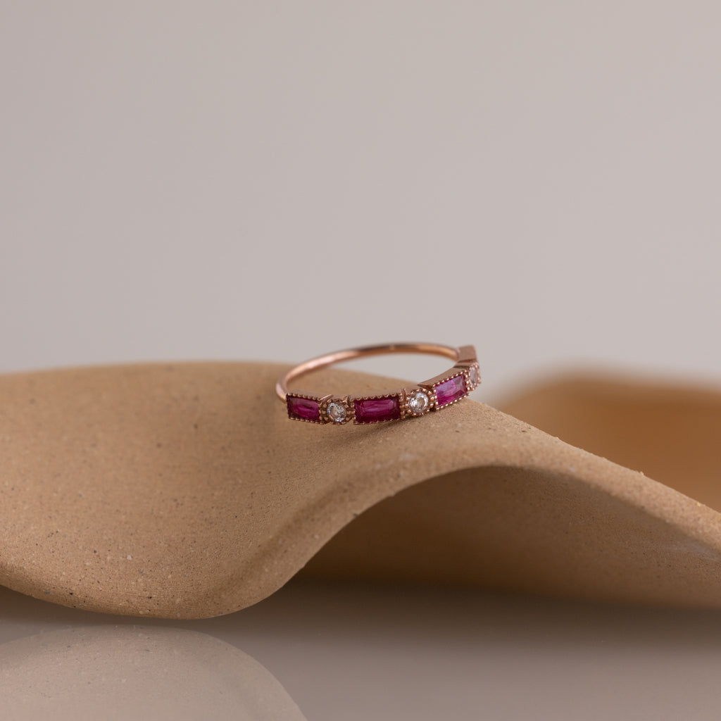 The Mixed Art Deco Birthstone Ring in Rose Gold, featuring pink stones and small diamonds, is displayed on a curved beige surface.