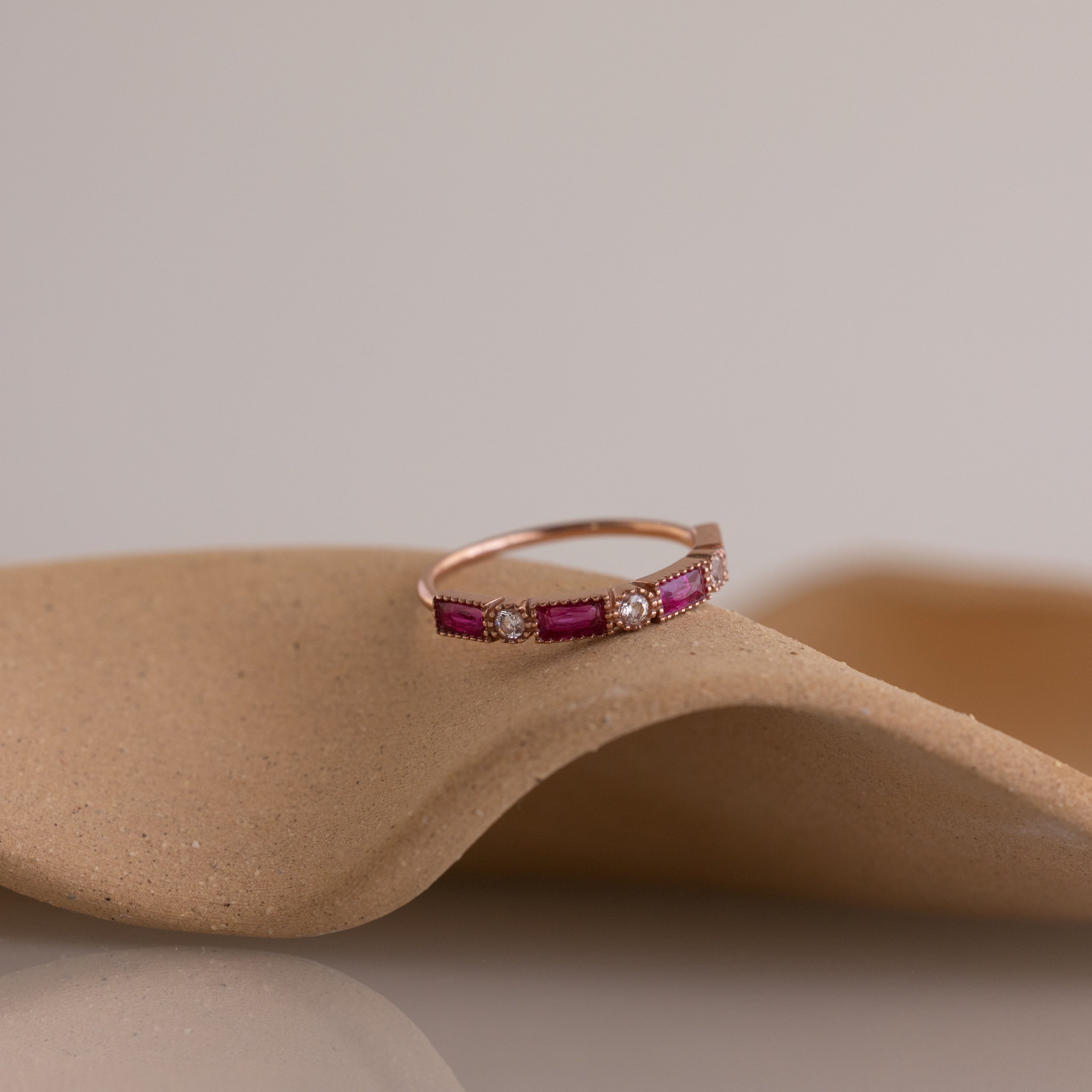 The Mixed Art Deco Birthstone Ring in Rose Gold, featuring pink stones and small diamonds, is displayed on a curved beige surface.