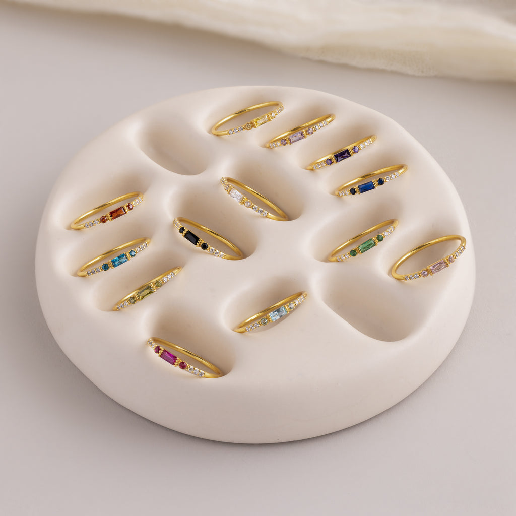 A full collection of dainty gold birthstone rings arranged neatly in a soft cream ceramic ring holder. Each ring features a rectangular baguette-cut gemstone in a range of vibrant colors—red, blue, green, pink, black, and more—flanked by small clear accent stones along the thin gold band.