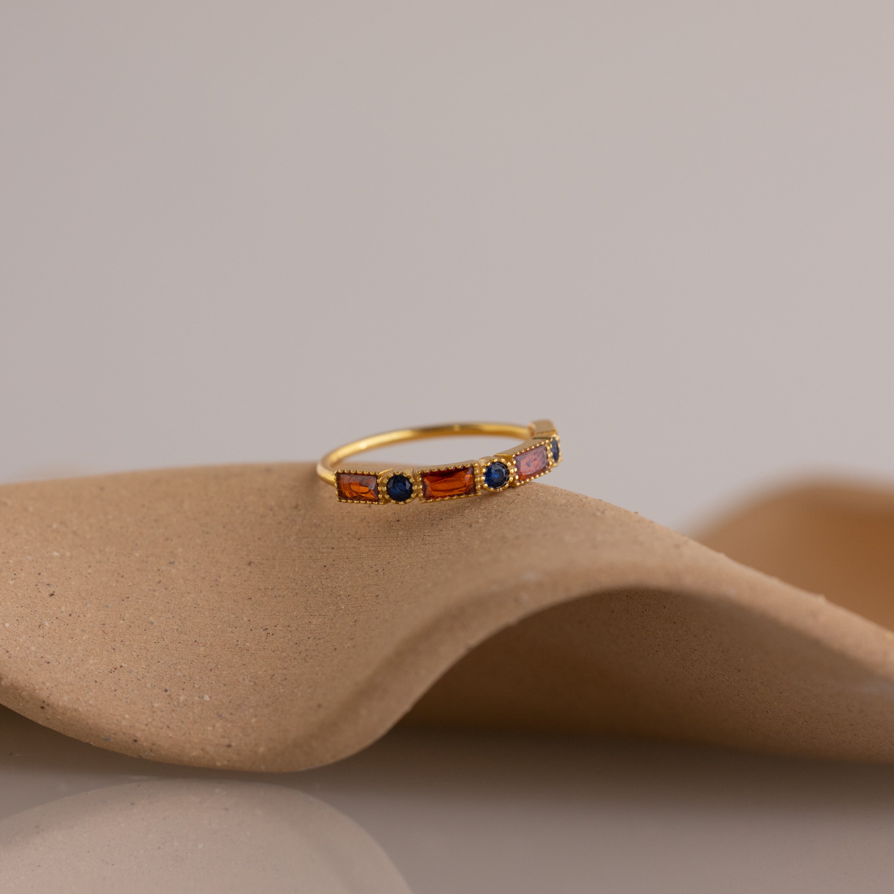 The Mixed Art Deco Birthstone Ring in 18K Gold, featuring vibrant orange and blue stones, is displayed on a textured beige surface, highlighting its bold colors and timeless design.