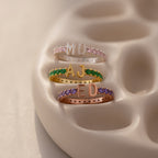 A Duo Initial Birthstone Tennis Ring in Amethyst (February), customized with letters MO, AJ, and ED, each featuring vibrant gemstone bands and displayed on a beige holder.