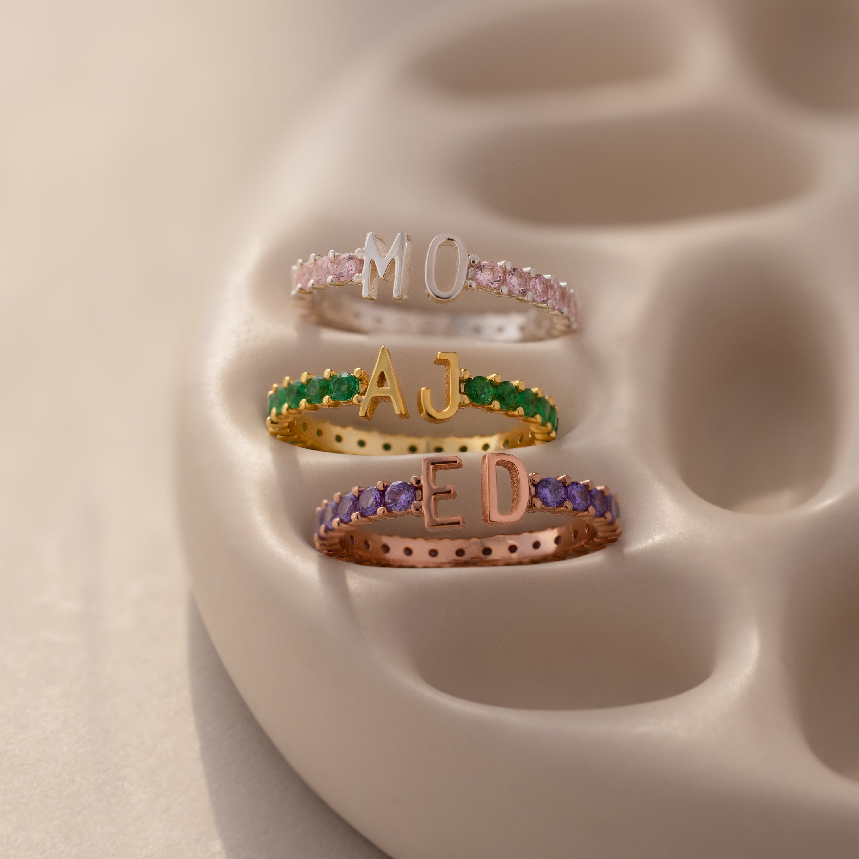 A Duo Initial Birthstone Tennis Ring in Amethyst (February), customized with letters MO, AJ, and ED, each featuring vibrant gemstone bands and displayed on a beige holder.