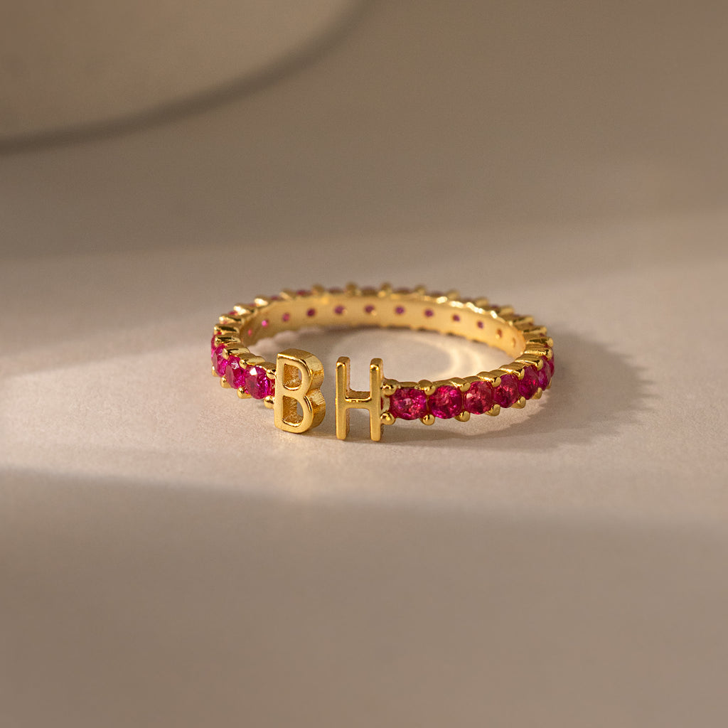 The Duo Initial Birthstone Tennis Ring in Garnet (January) features custom letters B and H on a neutral surface—a striking personalized jewelry piece for a unique touch.