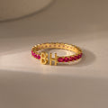 The Duo Initial Birthstone Tennis Ring in Garnet (January) features custom letters B and H on a neutral surface—a striking personalized jewelry piece for a unique touch.