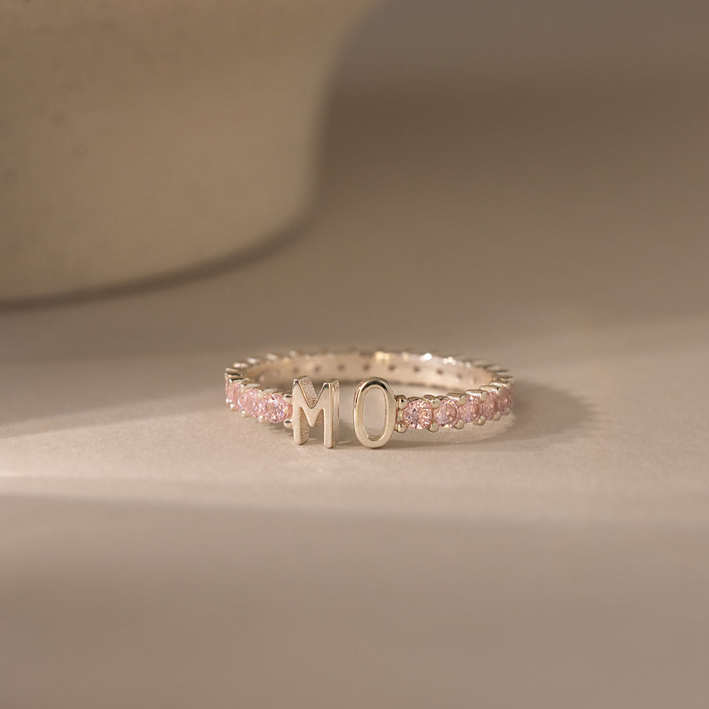 The Duo Initial Pink Tourmaline Tennis Ring features sparkling pink tourmaline gemstones and customizable letters M and O, displayed on a neutral surface.