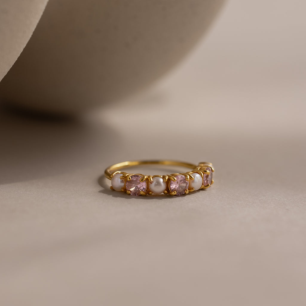 The Pink Tourmaline & Pearl Lace Ring features alternating white pearls and pink tourmaline gemstones set on a gold band, displayed on a soft beige surface.