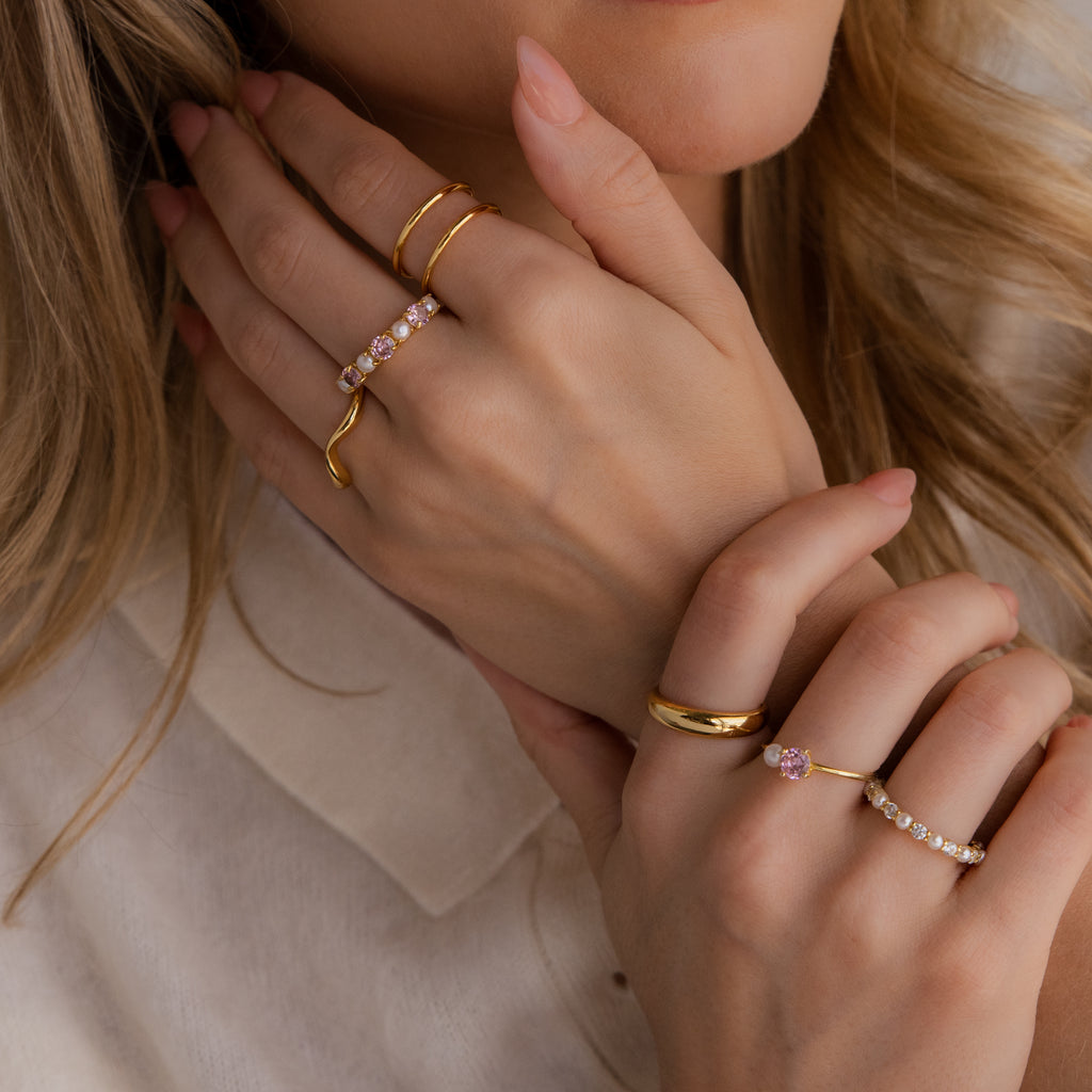 Close-up of a person wearing multiple gold rings, including the Pink Tourmaline & Pearl Lace Ring, featuring gemstones and diamonds on both hands near their face, dressed in a cream-colored top.