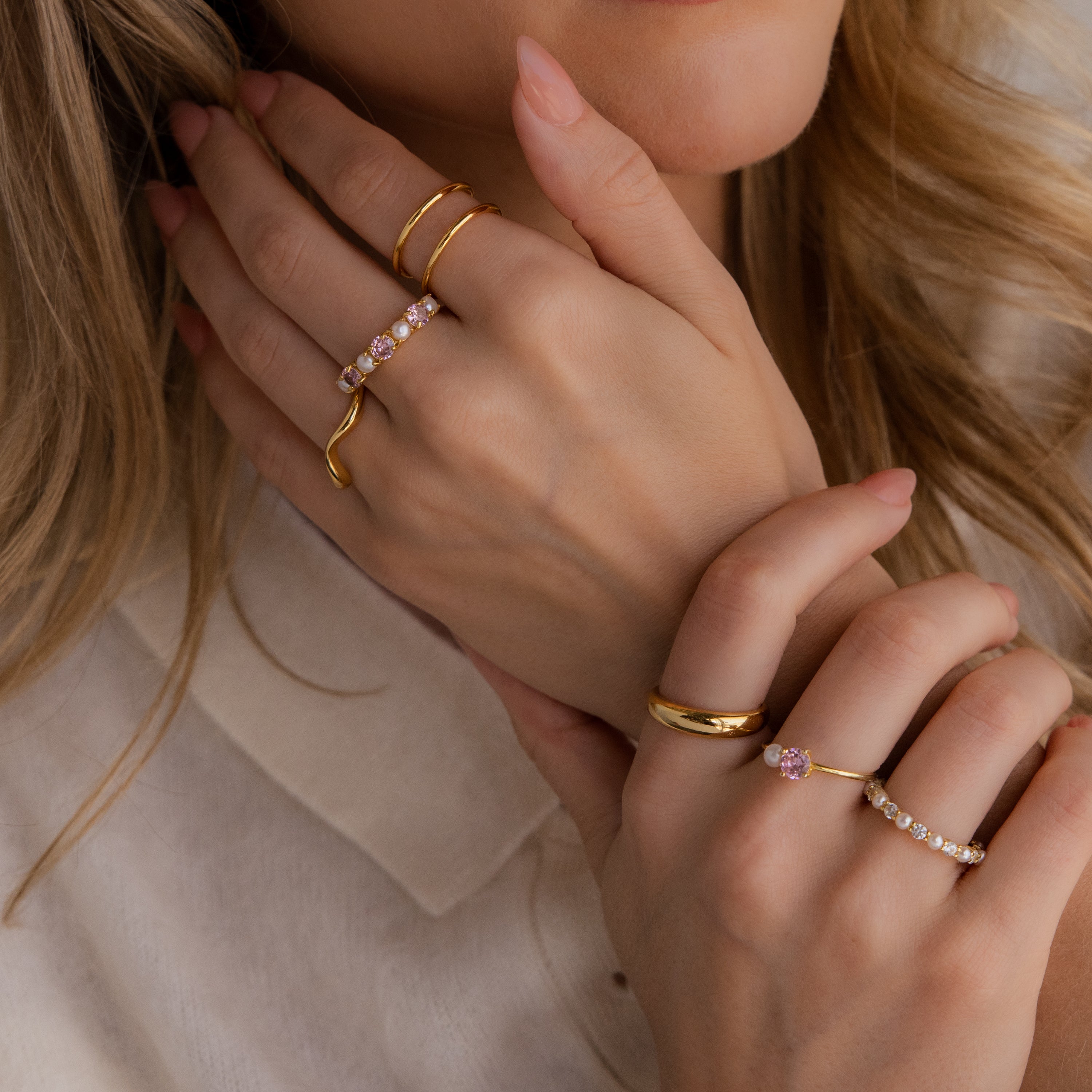 Close-up of a person wearing multiple gold rings, including the Pink Tourmaline & Pearl Lace Ring, featuring gemstones and diamonds on both hands near their face, dressed in a cream-colored top.