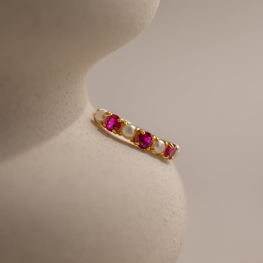 The Pink Ruby & Pearl Lace Ring, featuring alternating pink gemstones and pearls, rests on a smooth, curved beige surface.