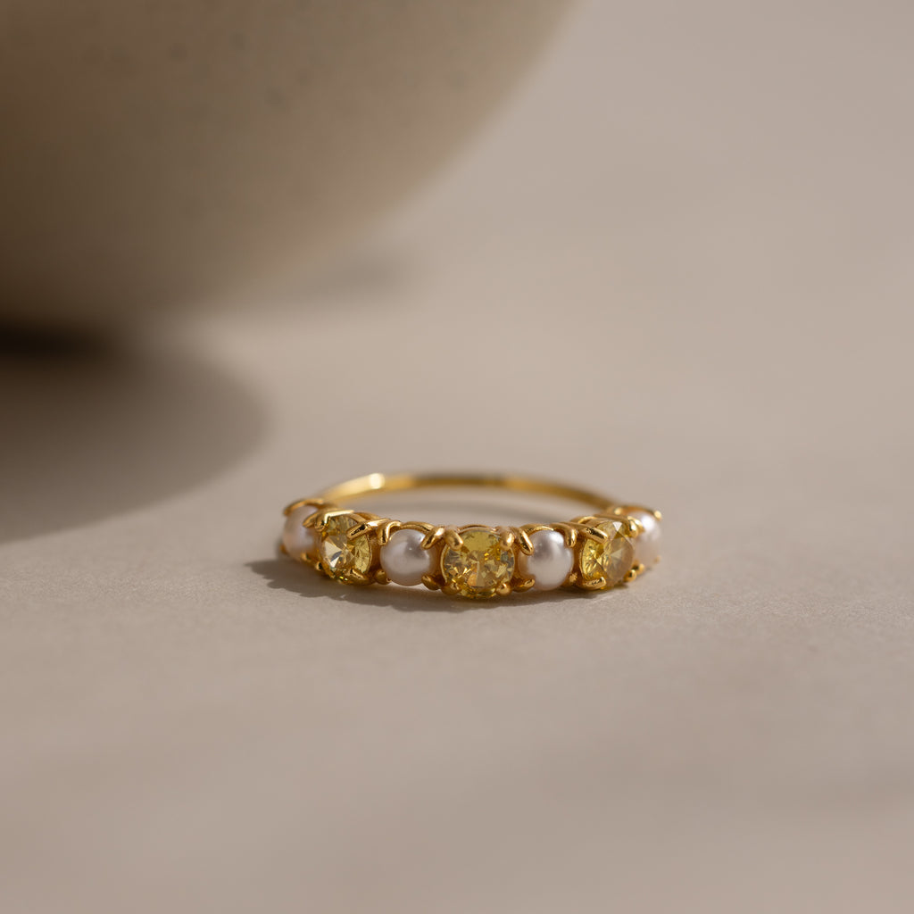 The Topaz & Pearl Lace Ring features a gold band adorned with yellow topaz gemstones and delicate white pearl-like beads, beautifully displayed on a neutral surface under soft lighting.