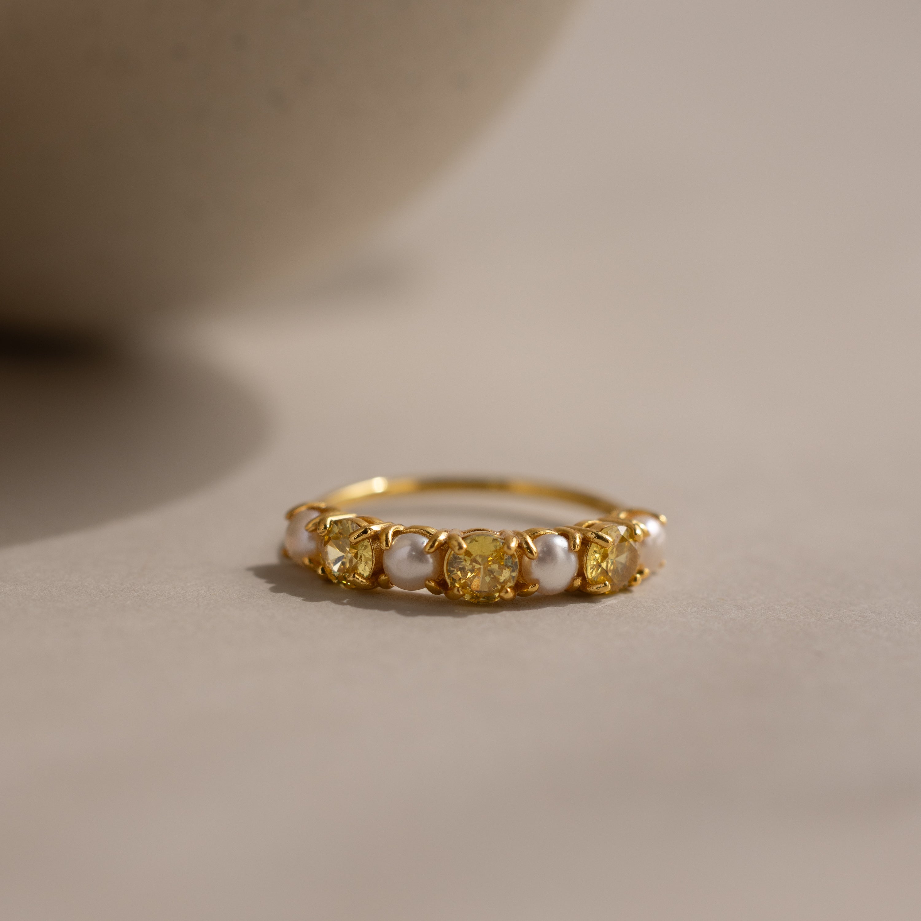 The Topaz & Pearl Lace Ring features a gold band adorned with yellow topaz gemstones and delicate white pearl-like beads, beautifully displayed on a neutral surface under soft lighting.