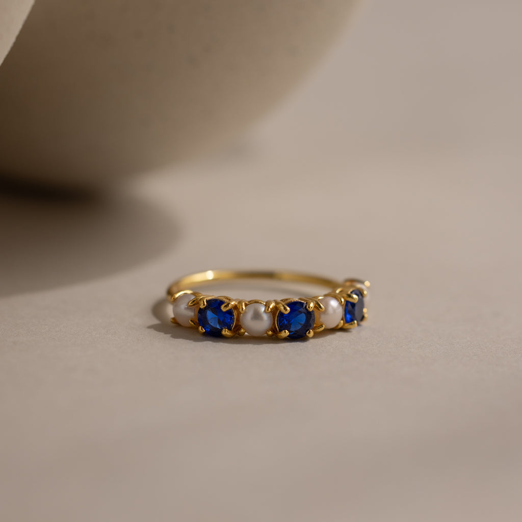 The Sapphire & Pearl Lace Ring features a lustrous pearl and blue sapphires set in a gold band, elegantly displayed on a neutral surface.