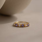 The Light Alexandrite & Pearl Lace Ring, featuring alternating pearls and shimmering light alexandrite stones in a gold band, rests elegantly on a beige surface.