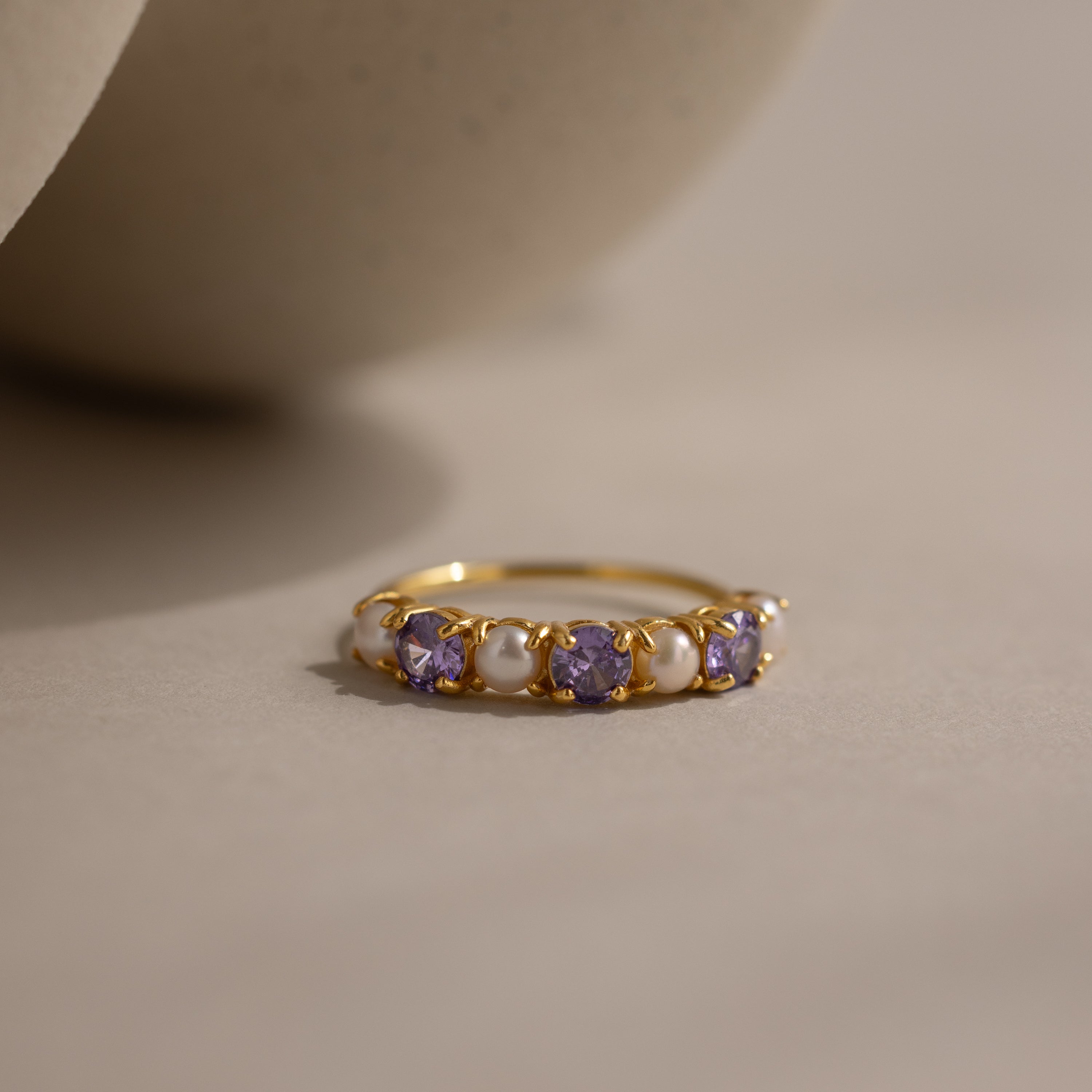 The Light Alexandrite & Pearl Lace Ring, featuring alternating pearls and shimmering light alexandrite stones in a gold band, rests elegantly on a beige surface.