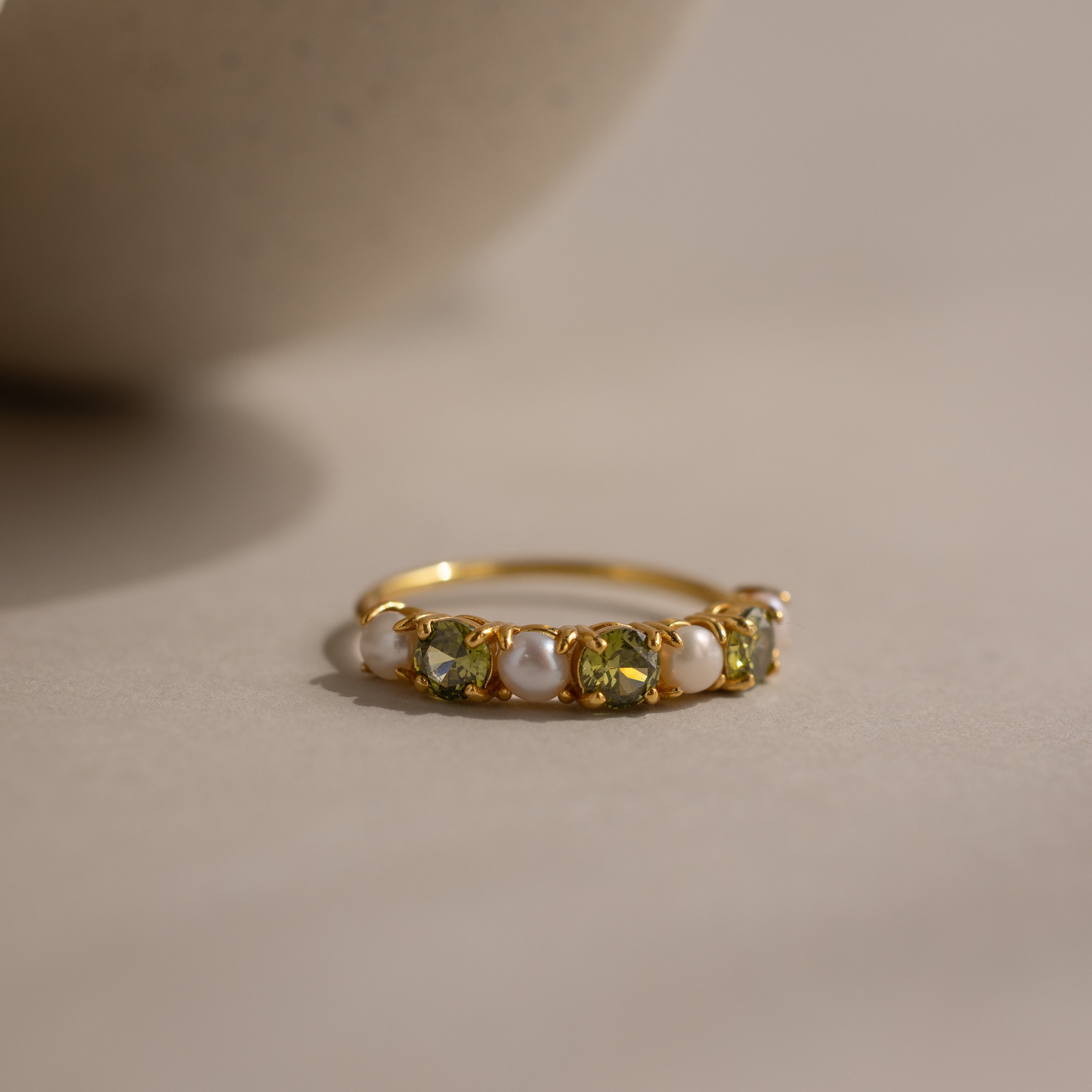 The Peridot & Pearl Lace Ring features a gold band with alternating green peridots and pearls, showcased on a neutral surface against a soft background.