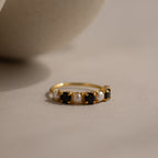 The Onyx & Pearl Lace Ring, a gold band with alternating black onyx and white pearl gemstones, is displayed on a neutral surface in soft lighting.